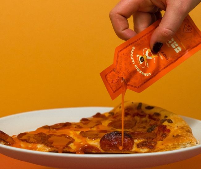 warm image of someone's hand, with black painted fingernailes, pouring infused hot sauce from an orange packet on to a slice of pizza, resting on a white plate