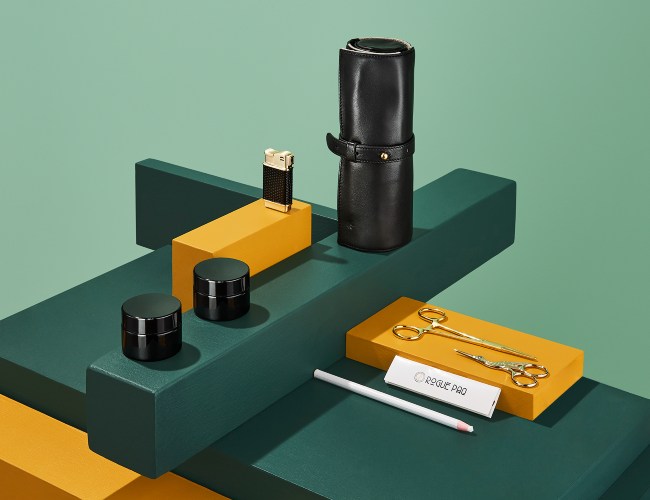 modern illustrated cubes, some elongated and some stout, some green and some yellow, provide a structure holding Rogue Paq's smoking accessories like a black leather pouch, black grinders, a black and gold lighter, gold scissors and white rolling papers