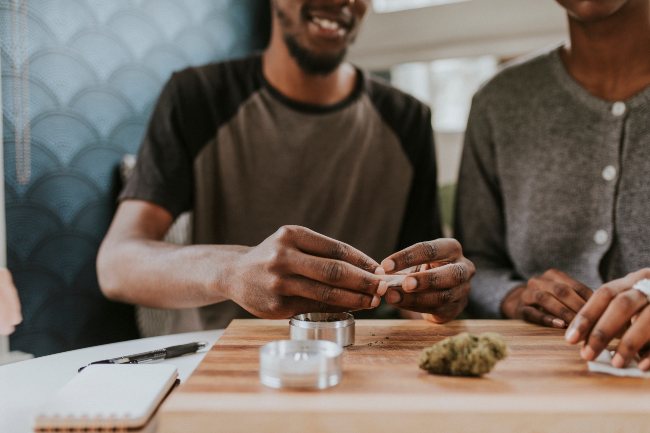 two people sitting down to rolling a joint with a weed nugget in front of them, a grinder, and rolling papers on top of a cutting board.