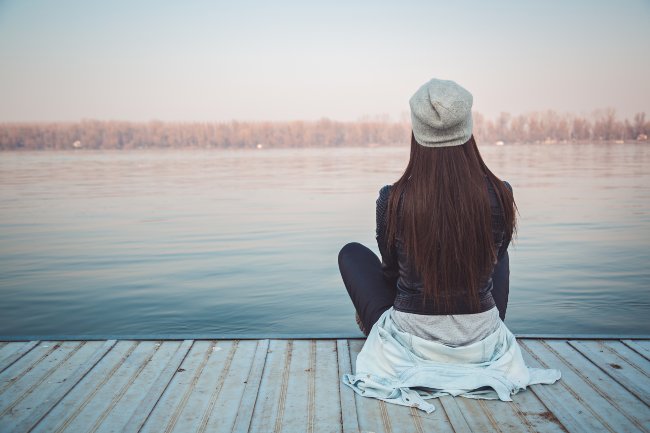 Women, with brown hair and hat on, sitting with her back turned towards photographer and she is looking out over a lake on a dock.