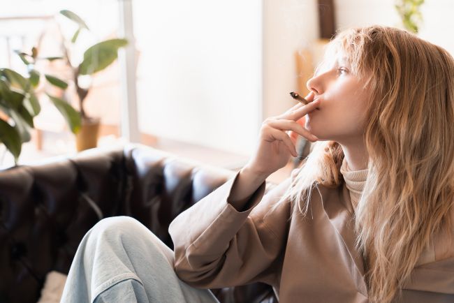 Women, with blonde hair and beige shirt, smoking a joint of cbd with smoke coming out