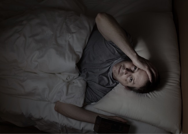 Man lying on his back with hand on his head, staring at the ceiling with blank eyes above him as lighting is dark.
