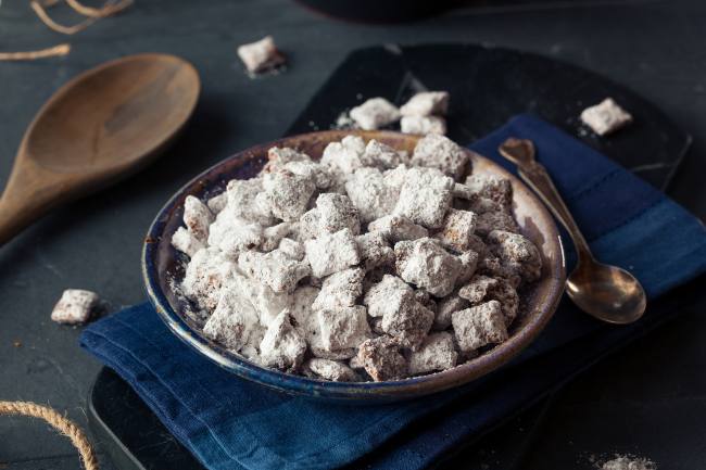 Bowl of muddy buddy mix that is composed of tiny squares covered in white powdered sugar that is lying on top of a navy blue towel on a grey counter with a brown wooden spoon next to it.