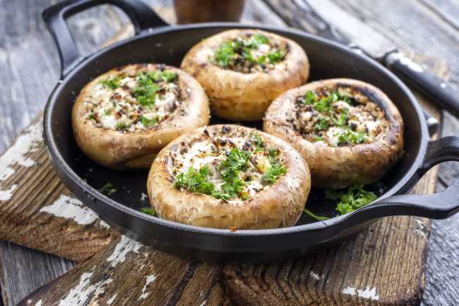 4 tan mushrooms lying in a cast iron skillet on top of a wooden board with white ricotta cheese and parsley inside the mushroos