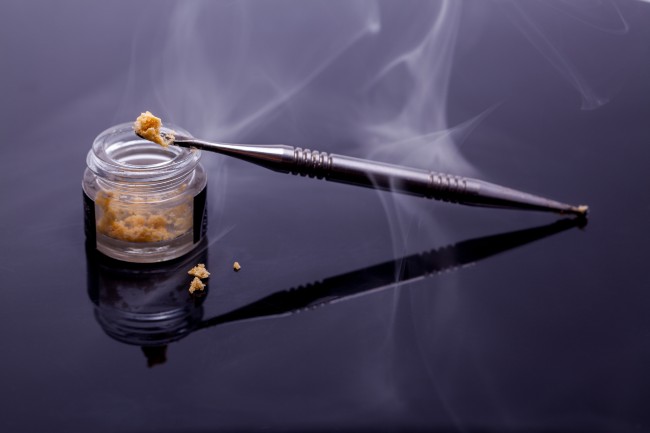 A jar of dabs on a black surface with a titanium dabber leaning against it and smoke over the top.