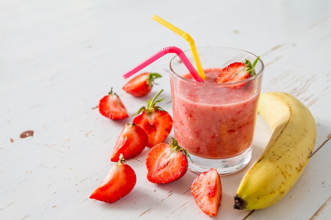 A straweberry banana smoothie in a class with straws and a banana and strawberries surrounding it