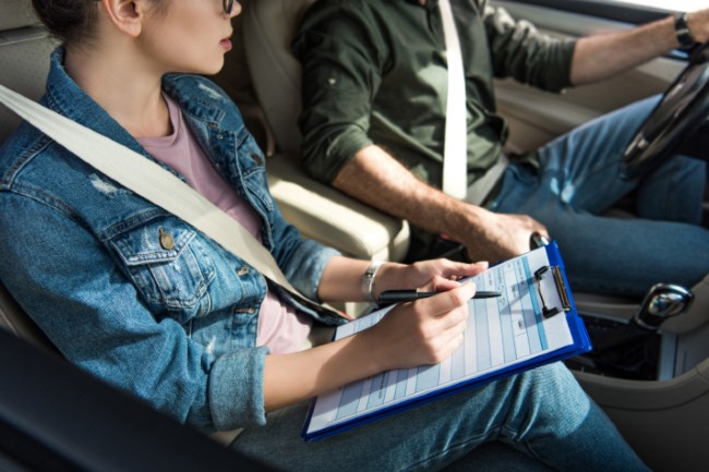 Two people sitting in a car. One is driving and one has a clipboard