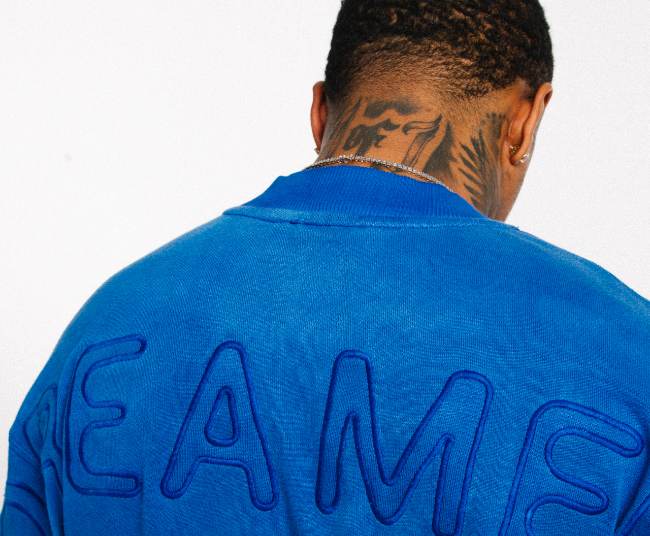 Man with a clean cut fade with his black hair as well as neck tattoos wearing a soft blue sweater with the word dreamers on the back.