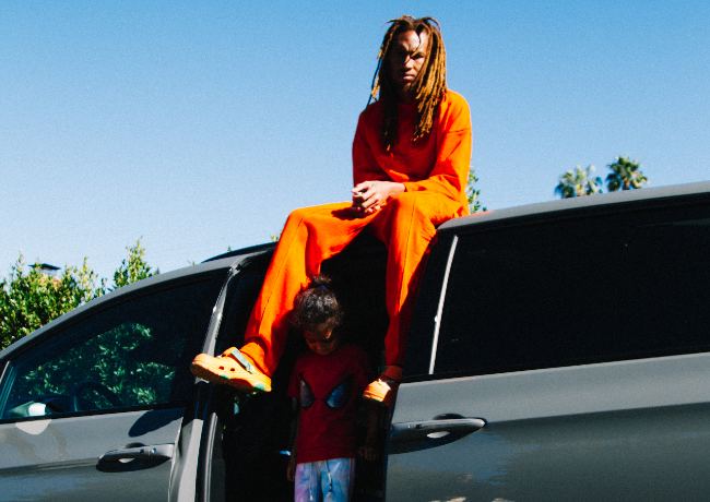 Black man with light brown dreadlocks sitting on top a van with the side door open and a small girl standing there. The man is wearing a Flowershop* orange sweater and orange sweatpants along with orange crocks.