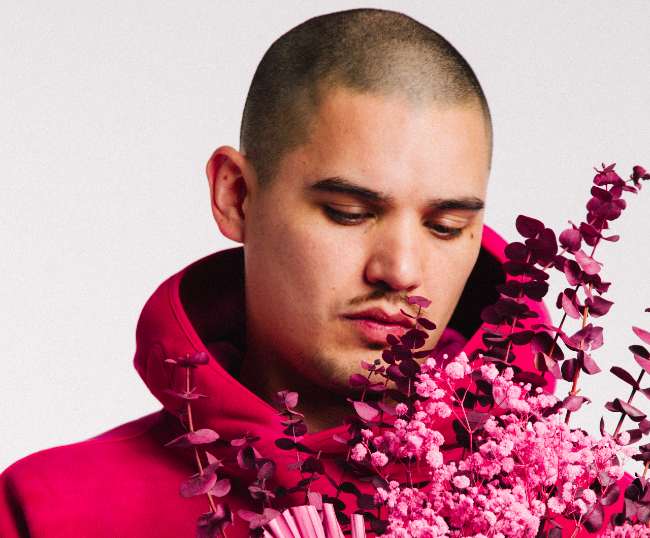 Man looking down at the pink and red flower bouquet he is holding while wearing a matching pink sweatshirt with hood.