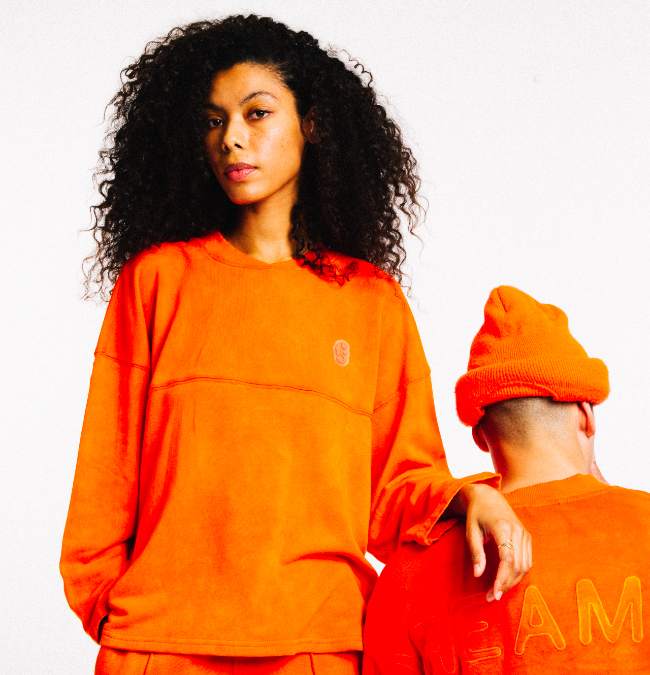 A woman stand while resting her arm on the shoulder of a man who is sitting down, with an orange hat on and hoodie to match the women's all orange outfit.