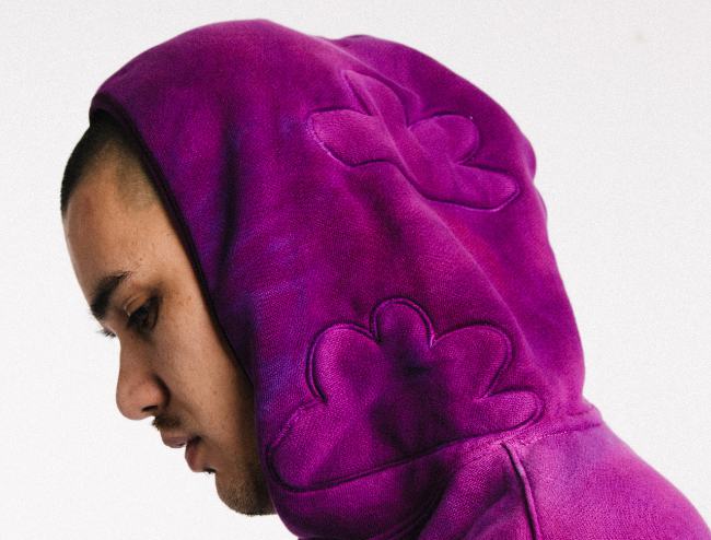 Man where a purple Flowershop* hoodie that has 2 clouds embossed on the hood that is pulled up above, around his head.