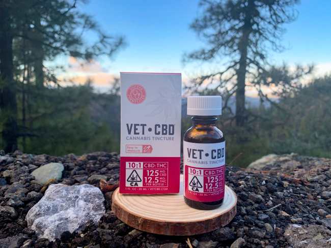 VETCBD tincture amber tincture bottle is being displayed with a white and red label and a white lid, next to its similar box packaging and a beautiful mountain view at sunset.