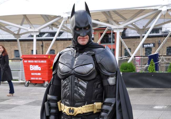 A man wearing a all black batman costume with pointed ears on a mask as well as a gold belt with a black belt on his waist, and a cape hanging off his back.