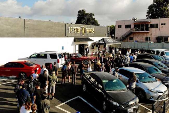 Image of Dr. Green Thumb's dispensary, that is decorated with a strip of black paint wrapped around the top of the building, with a pop-up tent in the corner of a parking lot with lots of cars and people in line to attend the event.