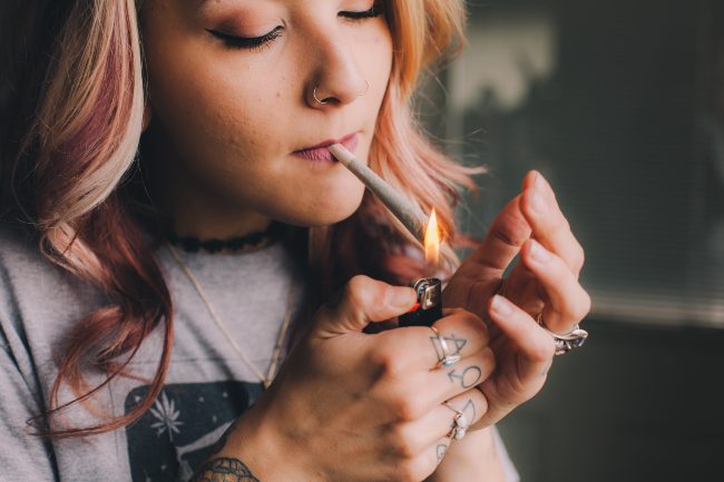 Women with pink and blonde hair that is curled with her hands cupping a joint as well as a lighting the joint with her one hand that has hand tattoos and rings.