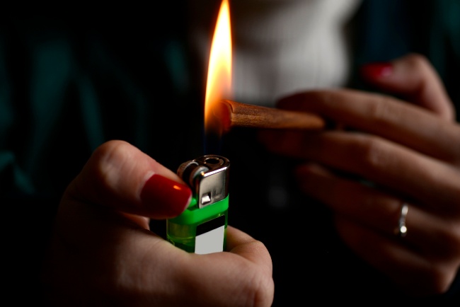 Woman's hand sparking a lighter by using her thumb, with red nail polish, to push down the button to produce a flame that is at the end of a blunt.