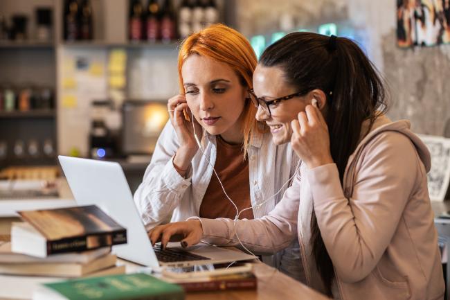 Two women staring at a laptop screen sharing a pair of earbud headphones with books surrounding them.