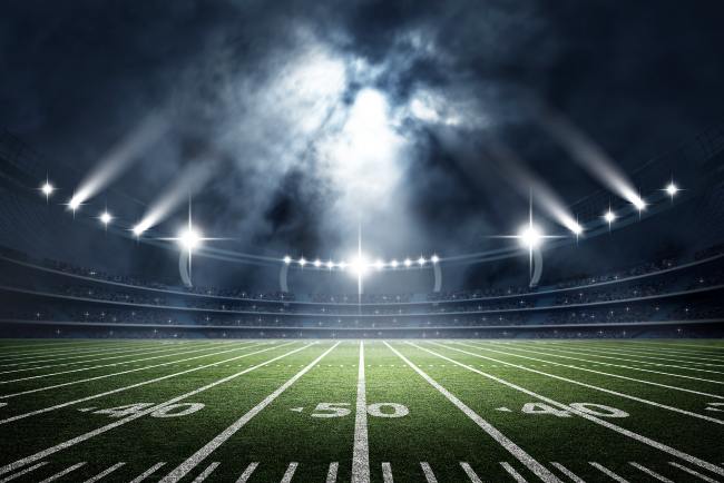 Image of a football stadium and lights shining down on the grass, painted lines, and painted numbers on the field with a smokey, dark ambience to the photograph.