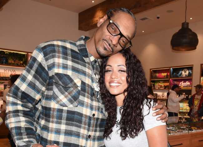 Snoop Dogg , wearing a plaid flannel, silver chain, and glasses, wrapping his arm around Tammy Pettigrew who is smiling with long dark and wavy curled hair.