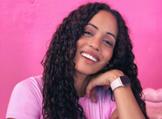 Black women smiling with bright white teeth and wavy long black hair with a Apple Watch on, wearing pink t-shirt and pink background.
