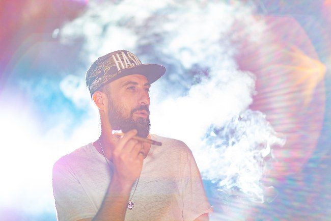 Man with beard and flat brim hat that says “HASH“ on the top with clouds iridescent lighting behind
            him and a weed blunt in his hand.