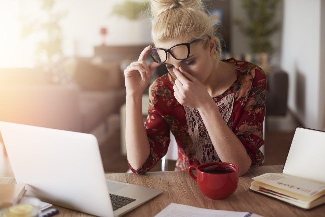 Une femme avec ses mains sur son visage dans la douleur, déplaçant ses lunettes sur son front tout en essayant de travailler sur son ordinateur portable à côté d'une tasse de café rouge.