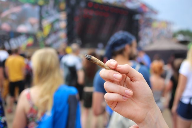 a woman’s hand holds up a joint at a music festival, with a stage in the background
