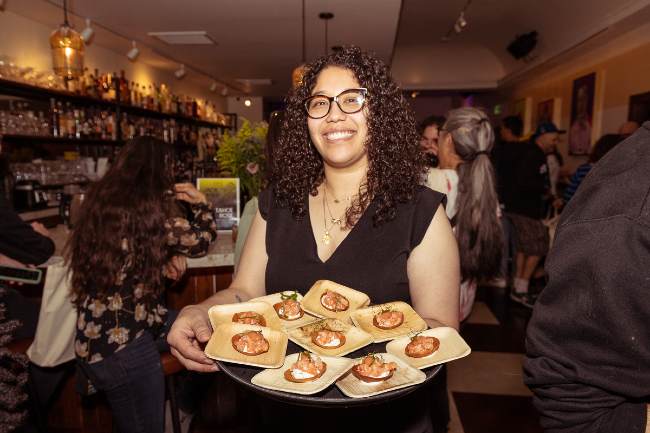 Woman standing with a platter of hors d'oeuvres on tan wooden plates, with white and orange food on top.