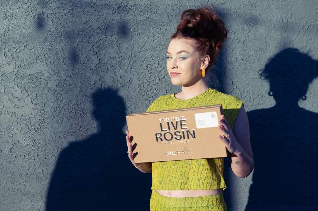 Lady in lime green outfit and red hair holding up a tan box that has “Live Resin“ on the front.
