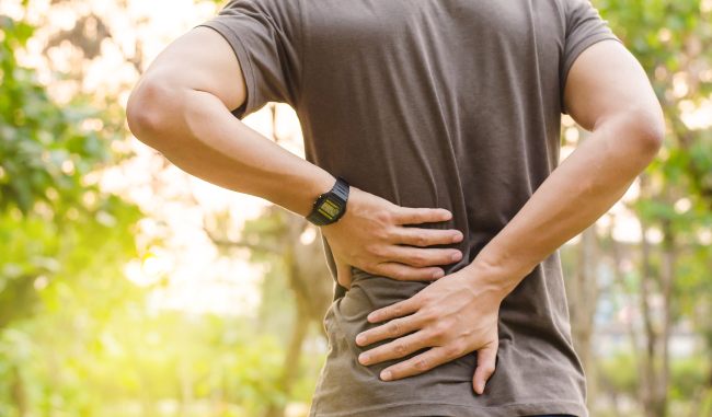 a man in a grey shirt, outside with trees in the background, grabs at his lower back like he is in pain