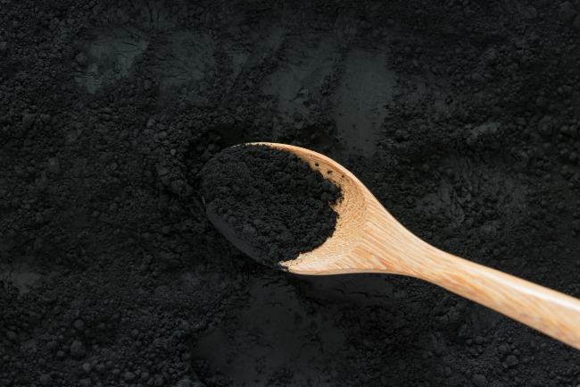 A wooden spoon scooping up activated charcoal from a black powder pile.