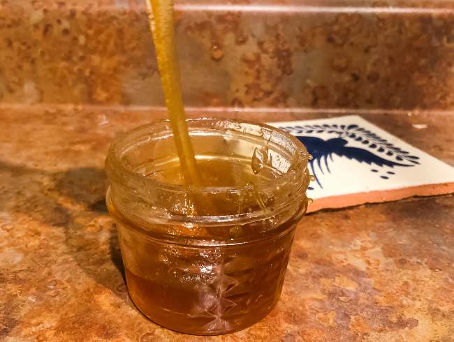 a thick golden strand of honey is pulled upwards from a small glass jar of homemade honey