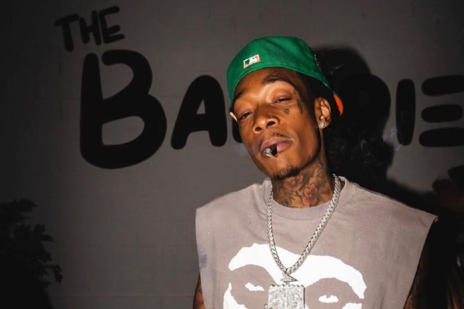 Whiz Khalifa with a cannabis joint in his mouth in front of a wall that says The Bakerie
