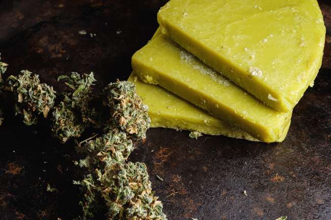 Three green slabs of cannabis butter lying on top of each other with a few dark green cannabis nugs beside it