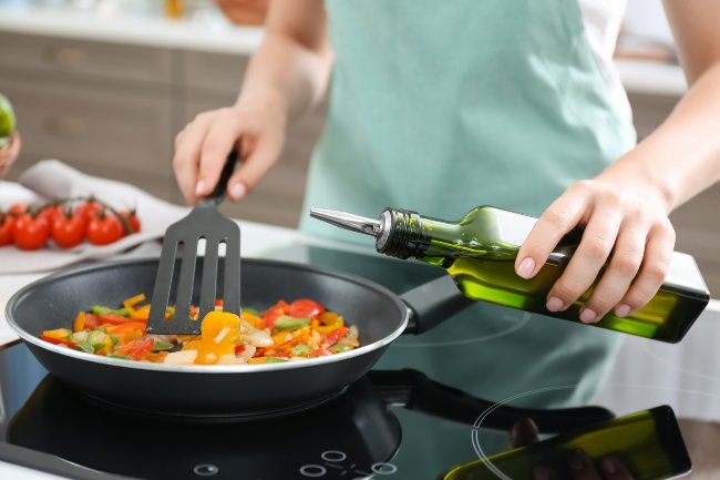 Person cooking a savory pasta in a black pan while pouring oil from a green oil bottle