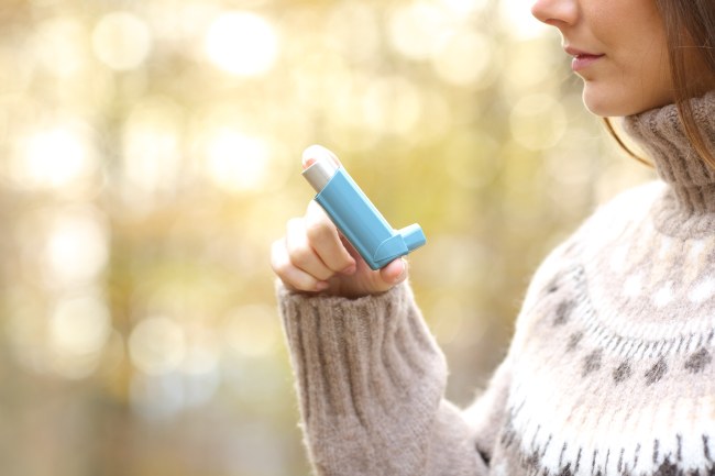 Woman holding a plastic, blue L-shaped inhaler to her mouth in the woods