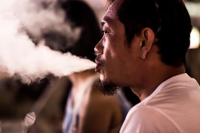 Profile view of a man blowing smoke out his mouth with people behind him that are out of focus