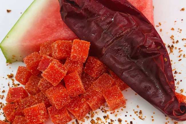 Red gummies and red salt lying next to a red chili pepper and red and green slice of watermelon.