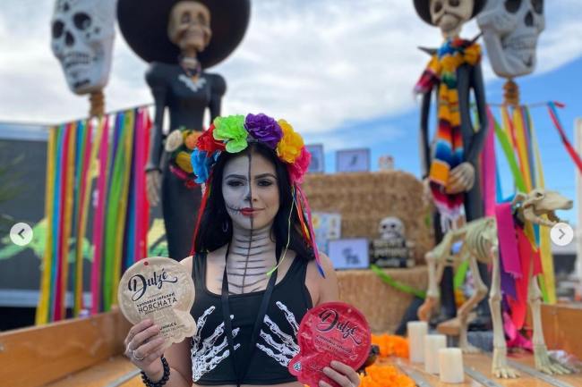 A young woman with a skull painted on her face holding a red DULZE Edibles package with skull decorations behind her