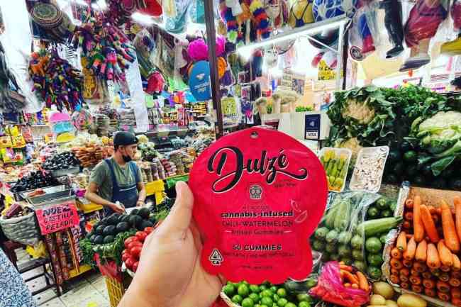 Hand holding a red package with DULZE written on it inside of a panderia with colorful piñatas hanging from the ceiling as well as fruits and vegetables on display for purchase