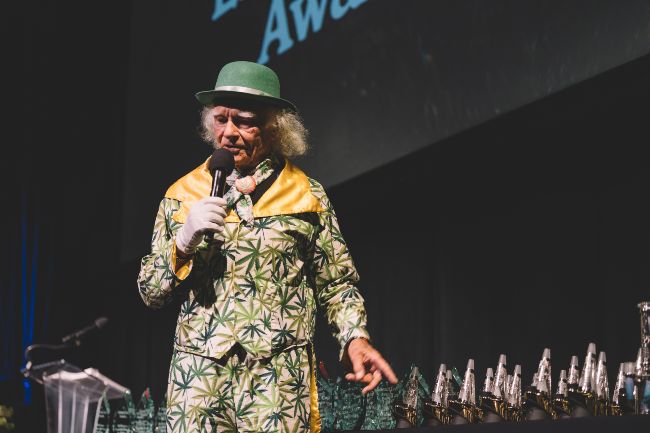 Man wearing a white and green marijuana leaf printed suit and a green hat, pointing at a bunch of Puffco awards.