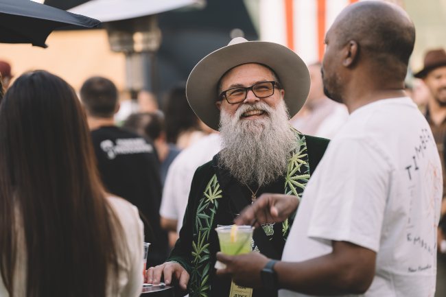 A man with a grey hat on, big grey beard, smiling, and wearing a vest with green cannabis leaves on it, talking to a black man with a neon green drink in hand