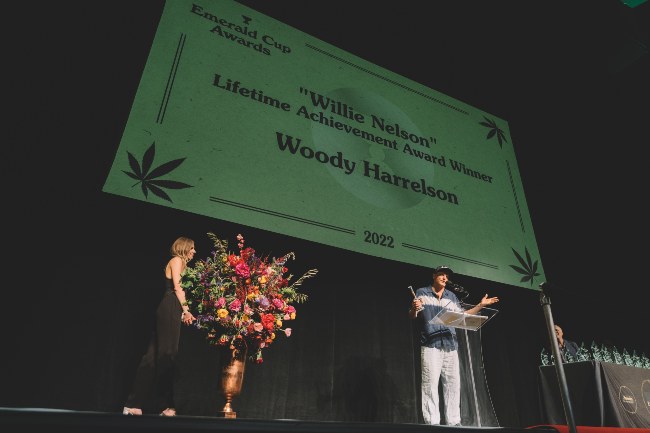 A giant, green sign with Woody Harrelson's name displayed with Woody Harrelson at the clear podium below on stage